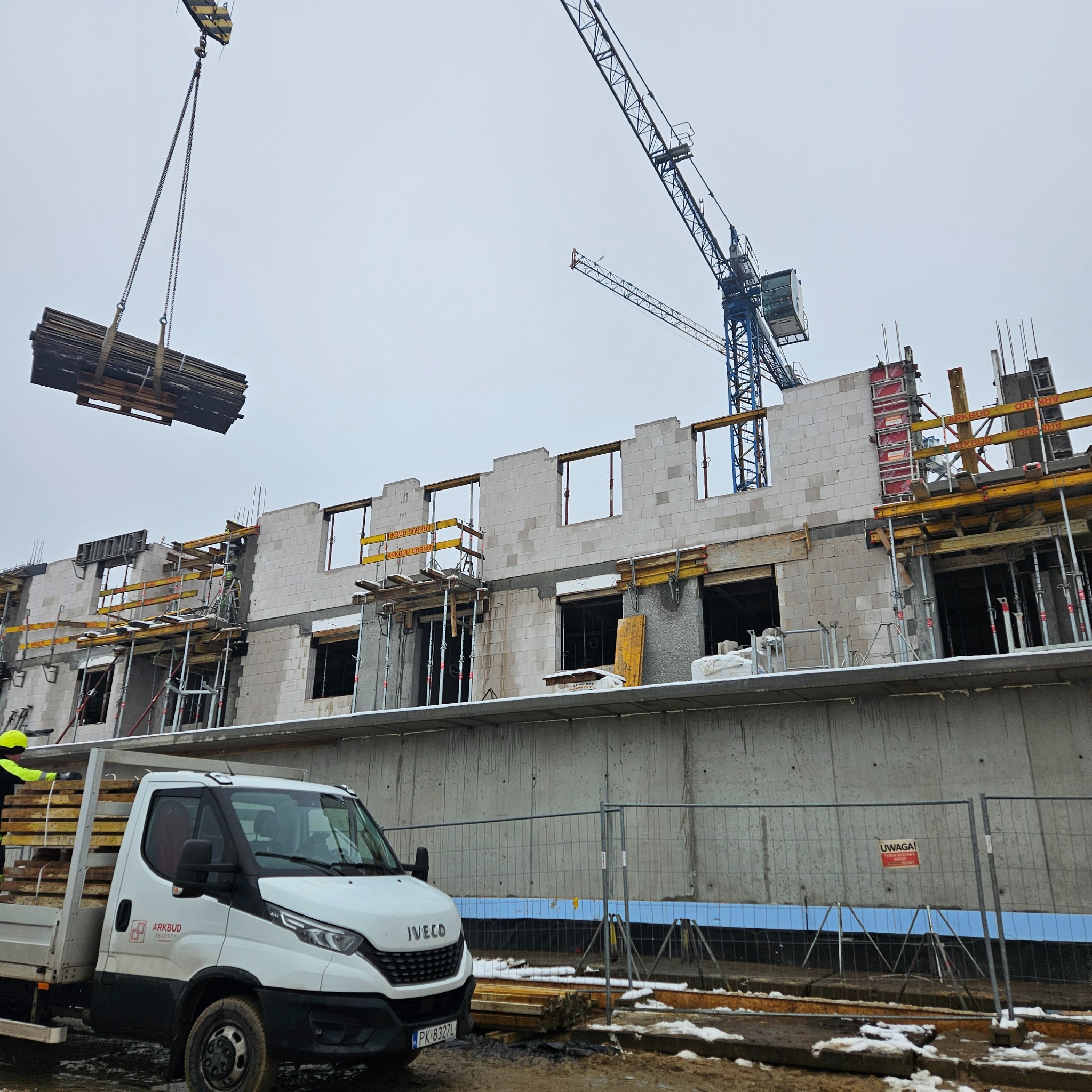 Budowa domu z betonu komórkowego: dźwig podnosi paletę z materiałami, widoczny fragment ściany, szalunki okienne i firmowy samochód dostawczy.