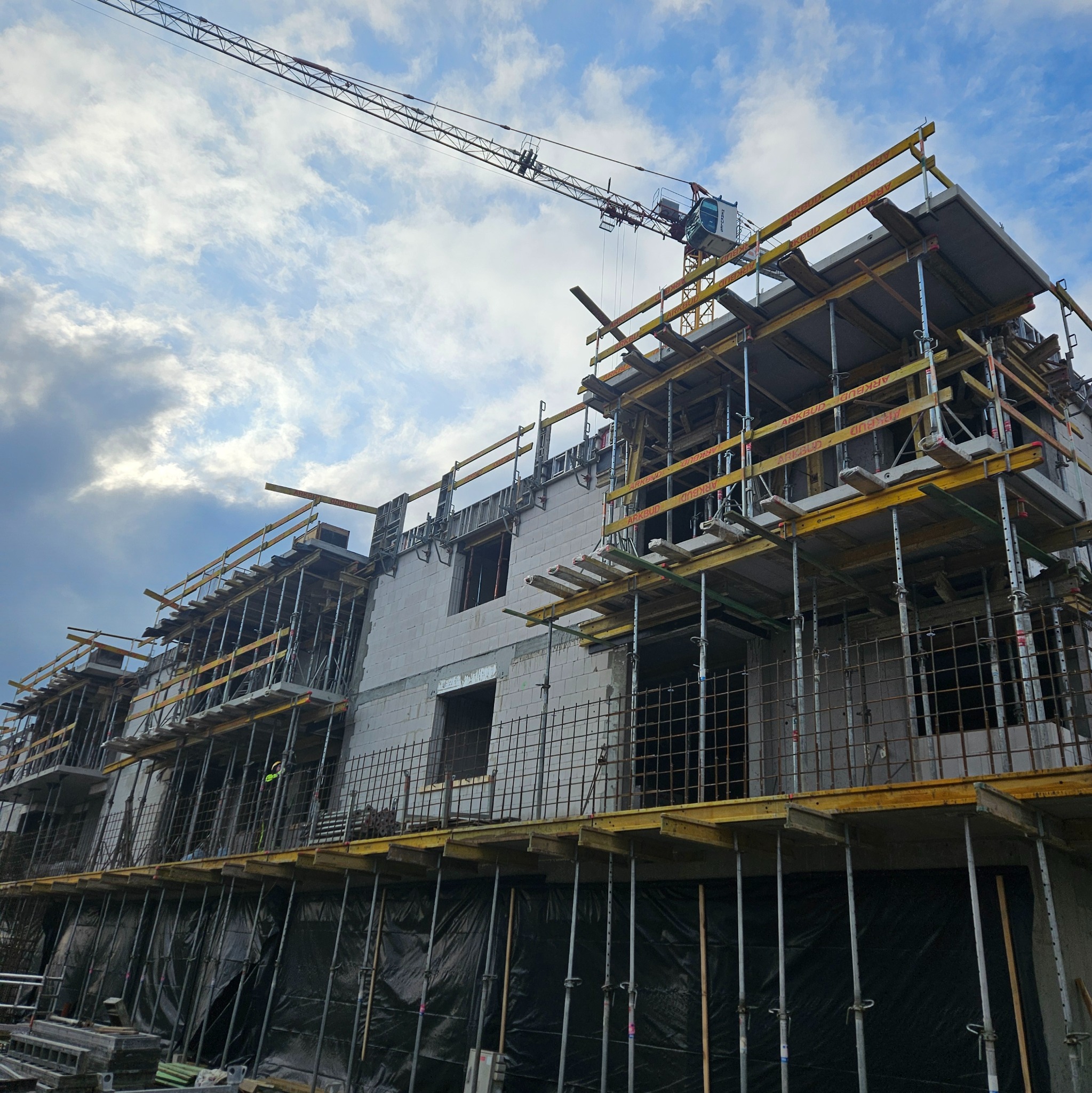Wznoszenie wielopiętrowego budynku z betonu komórkowego, widoczne rusztowania, podpory i żuraw budowlany na tle pochmurnego nieba. Etap murowania ścian.