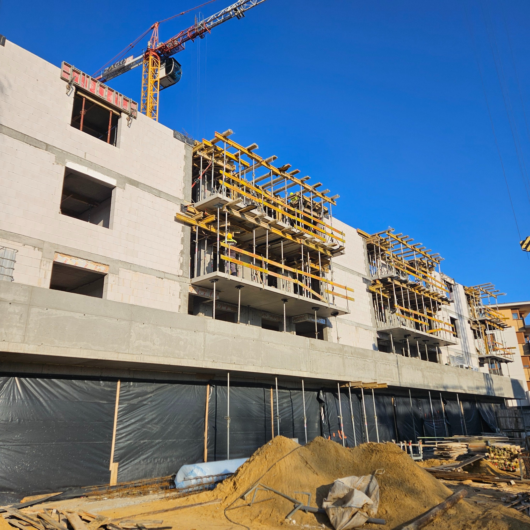 Budowa wielopiętrowego budynku z białego pustaka, widoczne szalunki balkonowe, żuraw budowlany w tle, piasek i materiały budowlane na pierwszym planie.