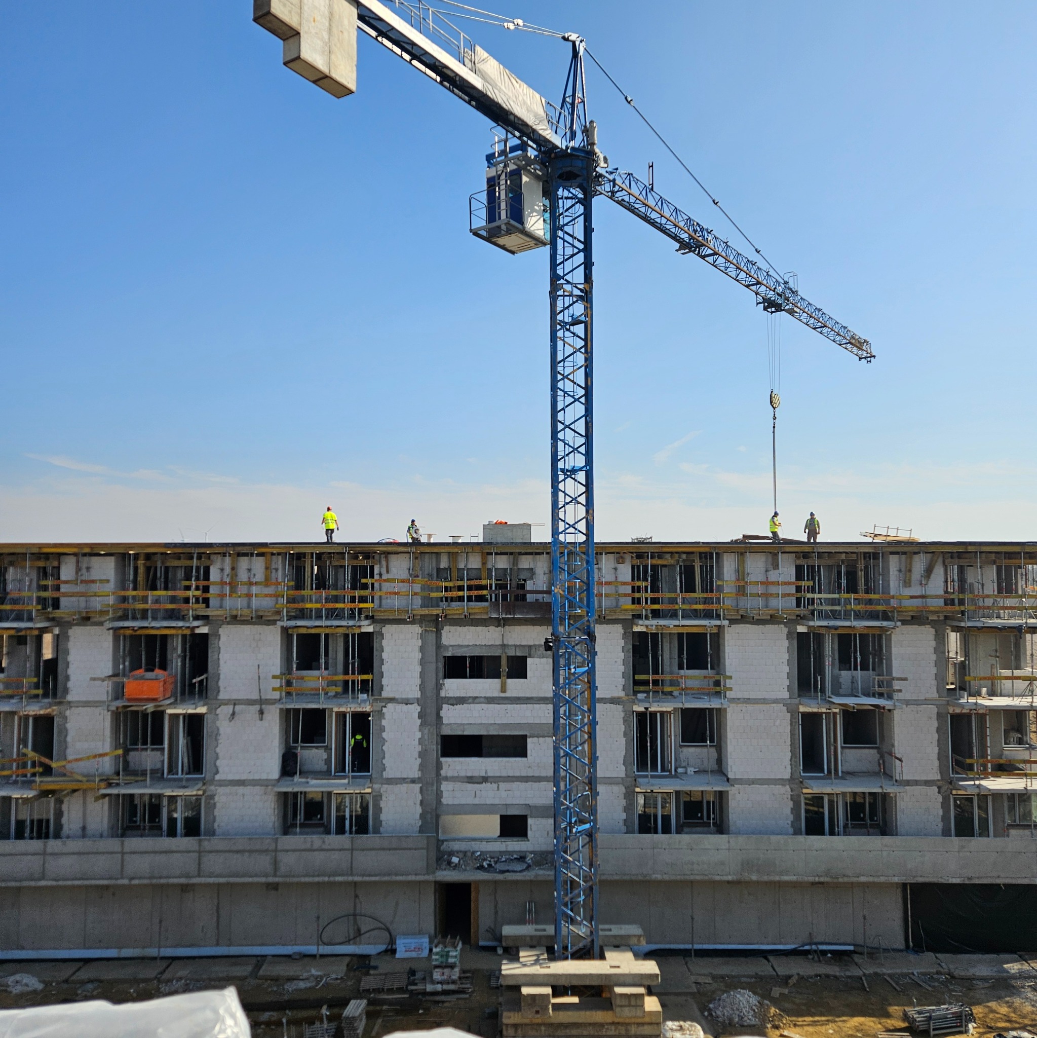 Wysoki żuraw budowlany na tle wznoszonego, wielopiętrowego budynku z betonu komórkowego. Widoczni pracownicy na górnej kondygnacji i szalunki stropowe.