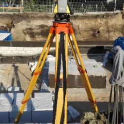 Żółty teodolit GeMax na trójnogu ustawiony na fundamencie z białych bloczków, w tle wykop, siatka ogrodzeniowa i budynek w stanie surowym otwartym.