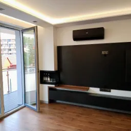 Nowoczesny salon z otwartym balkonem, drewnianą podłogą i czarną ścianą telewizyjną z minimalistyczną szafką RTV i półką na alkohol, oświetlony listwami LED na suficie.