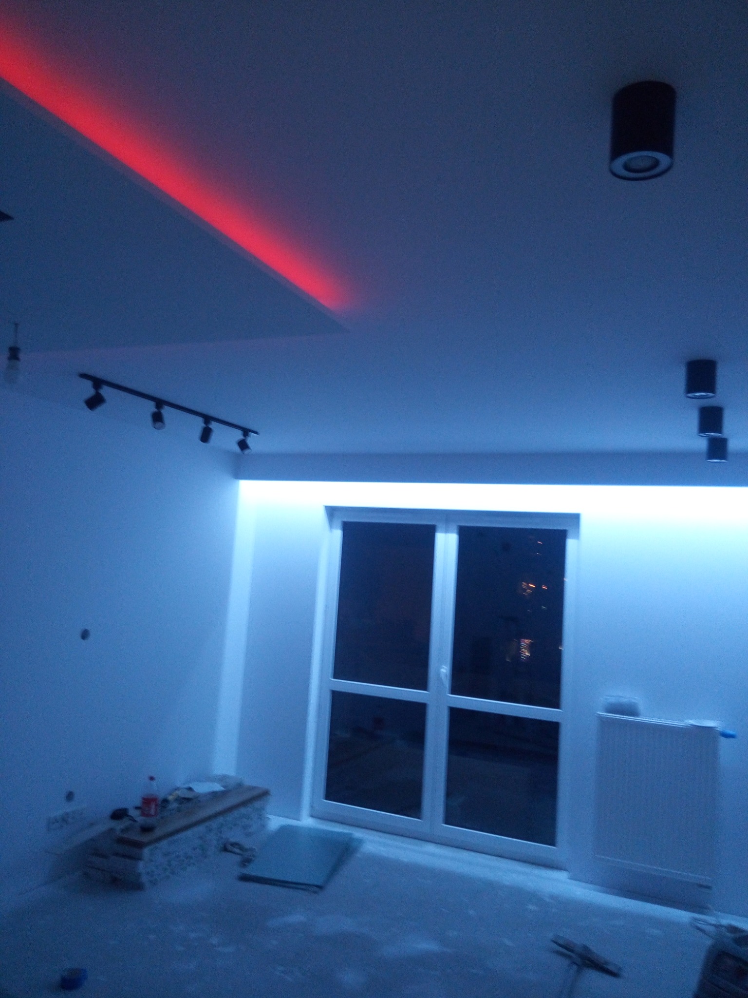 Pokój w trakcie remontu z podwieszanym sufitem z czerwonym oświetleniem LED i białym oświetleniem LED nad oknem balkonowym, widoczne narzędzia i materiały budowlane.