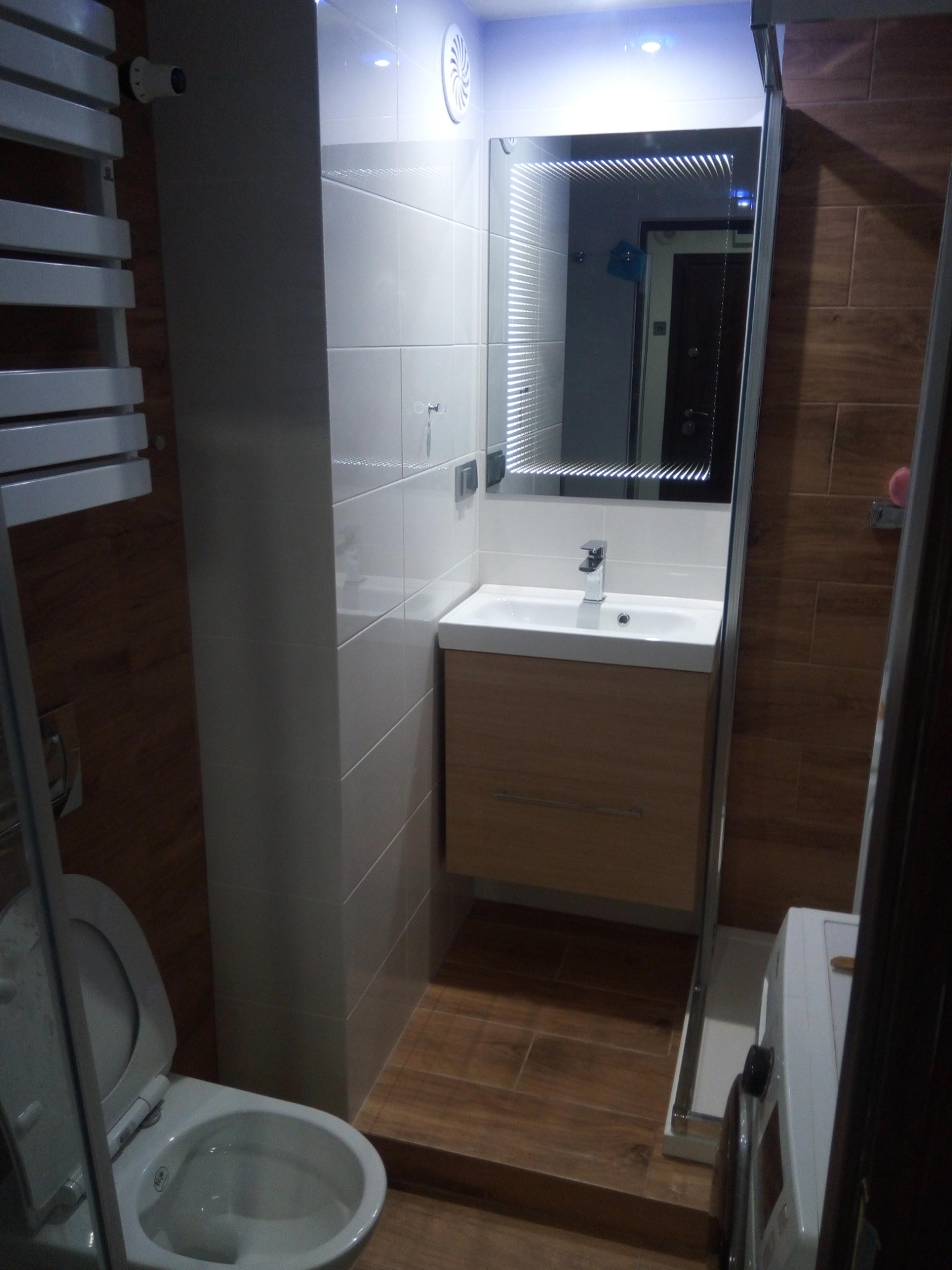 Nowoczesna łazienka z białymi i brązowymi płytkami, podświetlane lustro nad umywalką z szafką, grzejnik drabinkowy i toaleta z otwartą klapą.