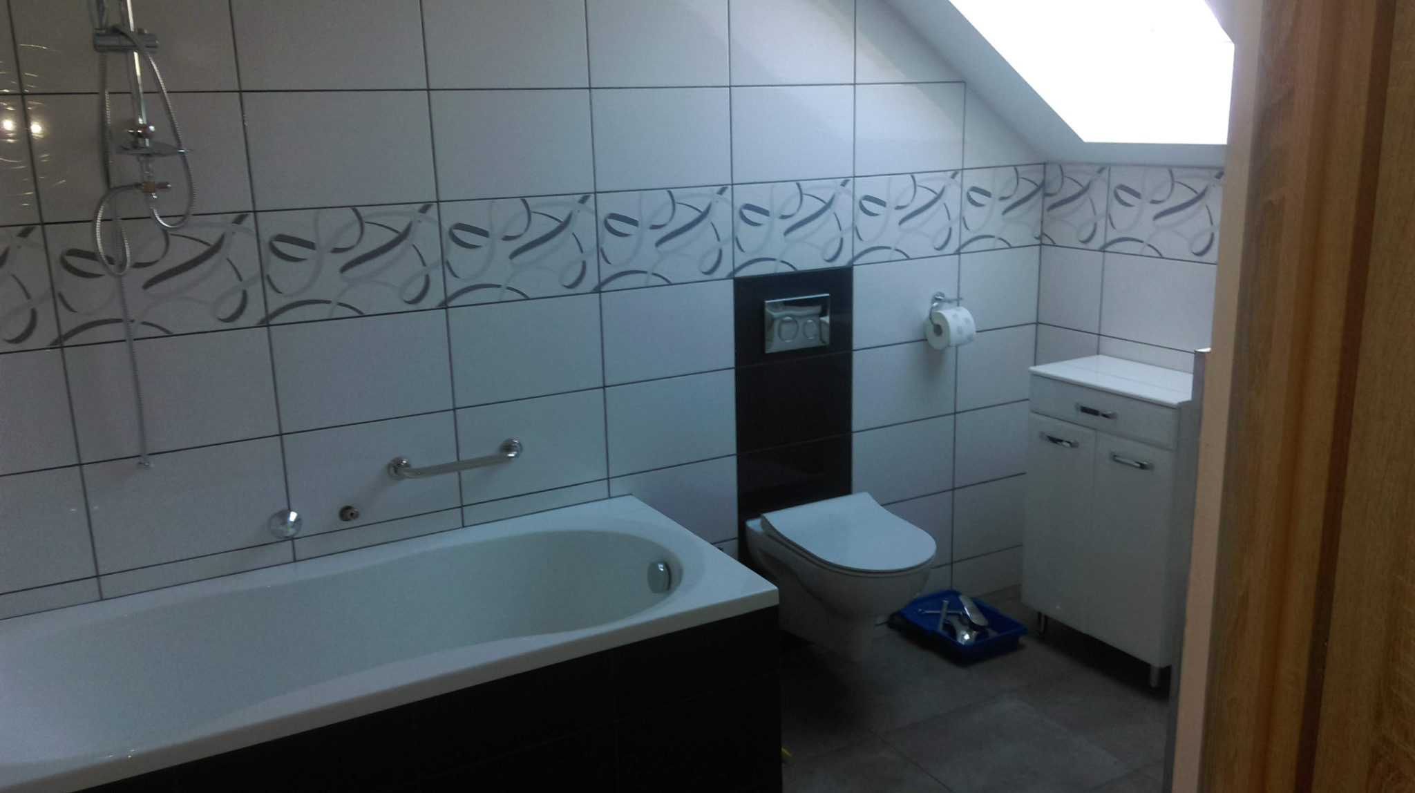 Wykończona łazienka na poddaszu z biało-czarnymi płytkami, wanną, toaletą podwieszaną i szafką pod umywalką, widoczne akcesoria łazienkowe i niebieska kuweta z narzędziami na podłodze.