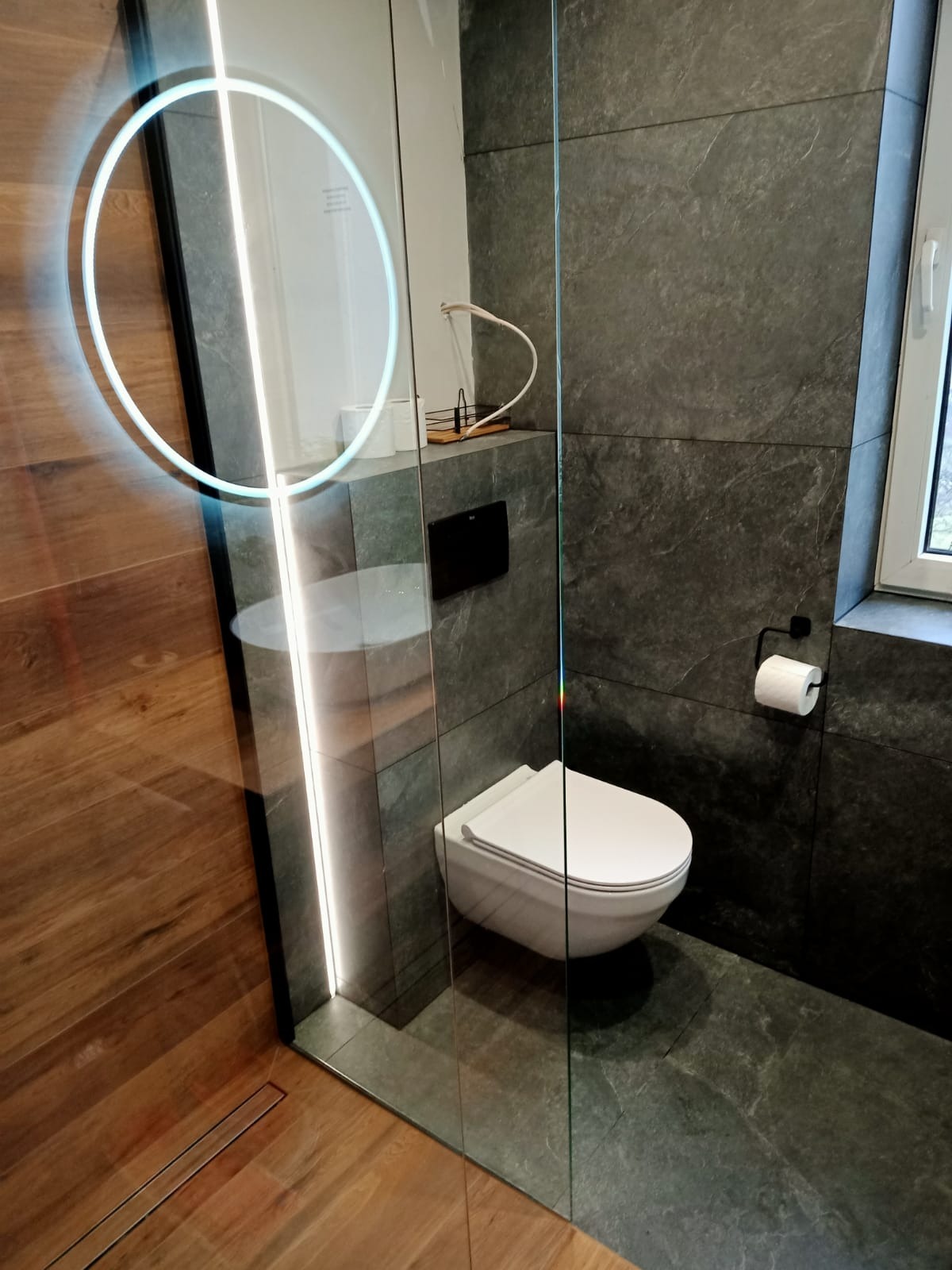 Nowoczesna łazienka z ciemnoszarą okładziną ścienną i podłogową, wiszącą toaletą oraz szklaną kabiną prysznicową z podświetlanym, okrągłym lustrem.