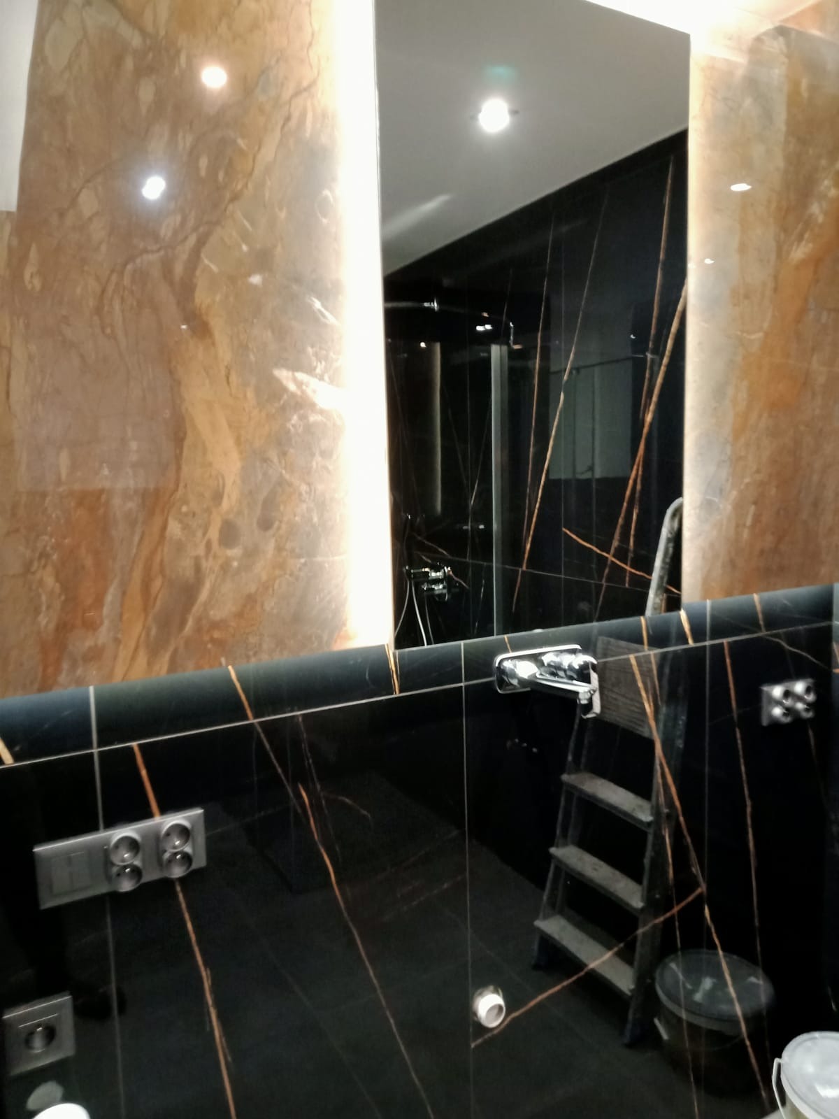 Luksusowa łazienka z czarnymi płytkami imitującymi marmur z złotymi żyłami, widoczne gniazdka elektryczne i drabina aluminiowa, odbicie kabiny prysznicowej w lustrze.