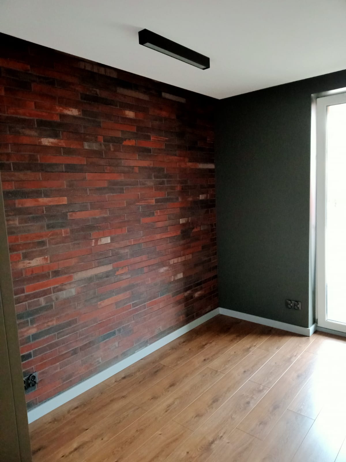 Pokój po remoncie z ceglaną ścianą, drewnianą podłogą i ciemnoszarą ścianą, oświetlony podłużną lampą sufitową.