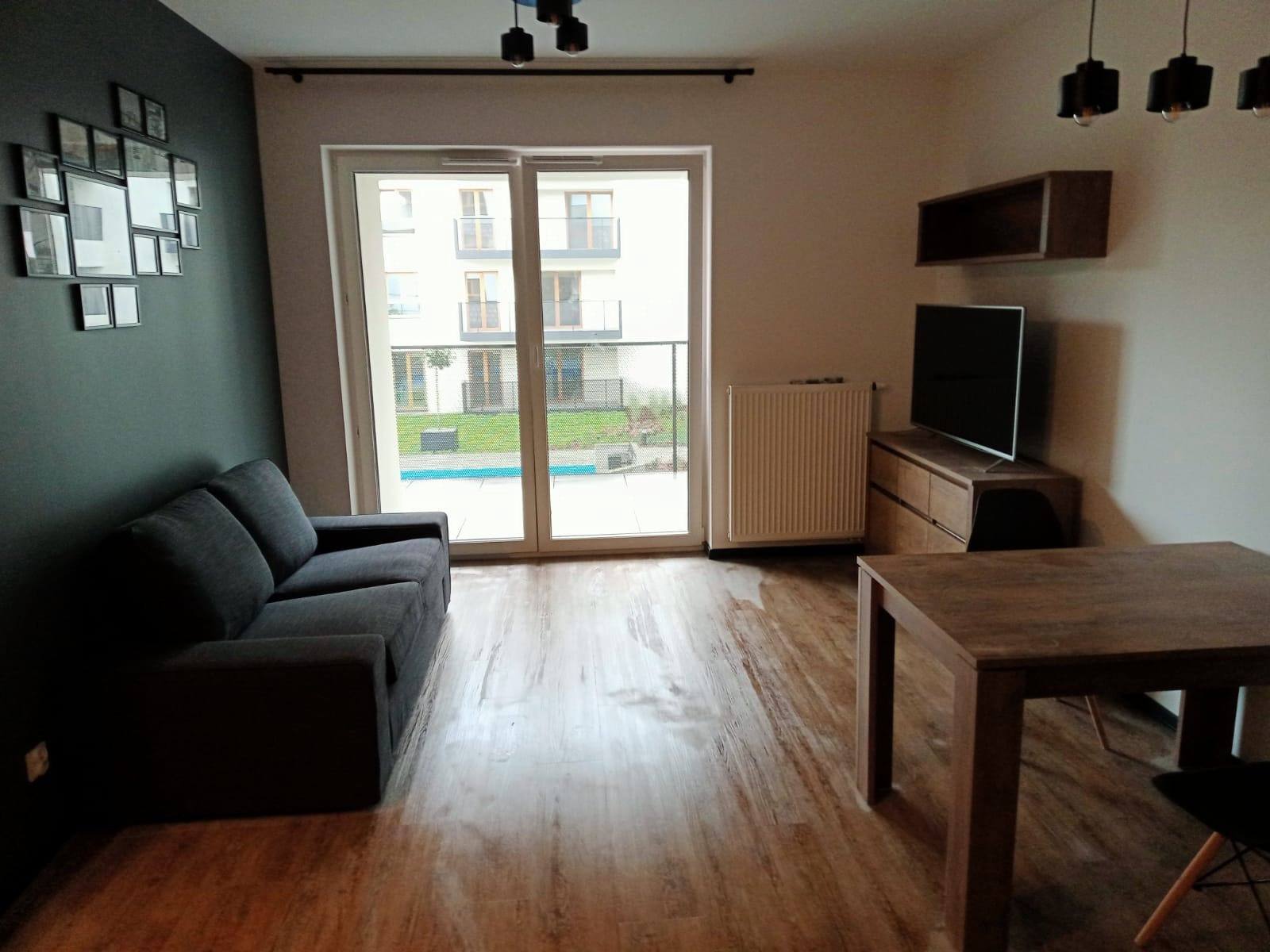 Nowoczesny salon z szarą sofą, drewnianym stołem i podłogą, dekoracją ścienną z ramek oraz widokiem na balkon przez duże okno.