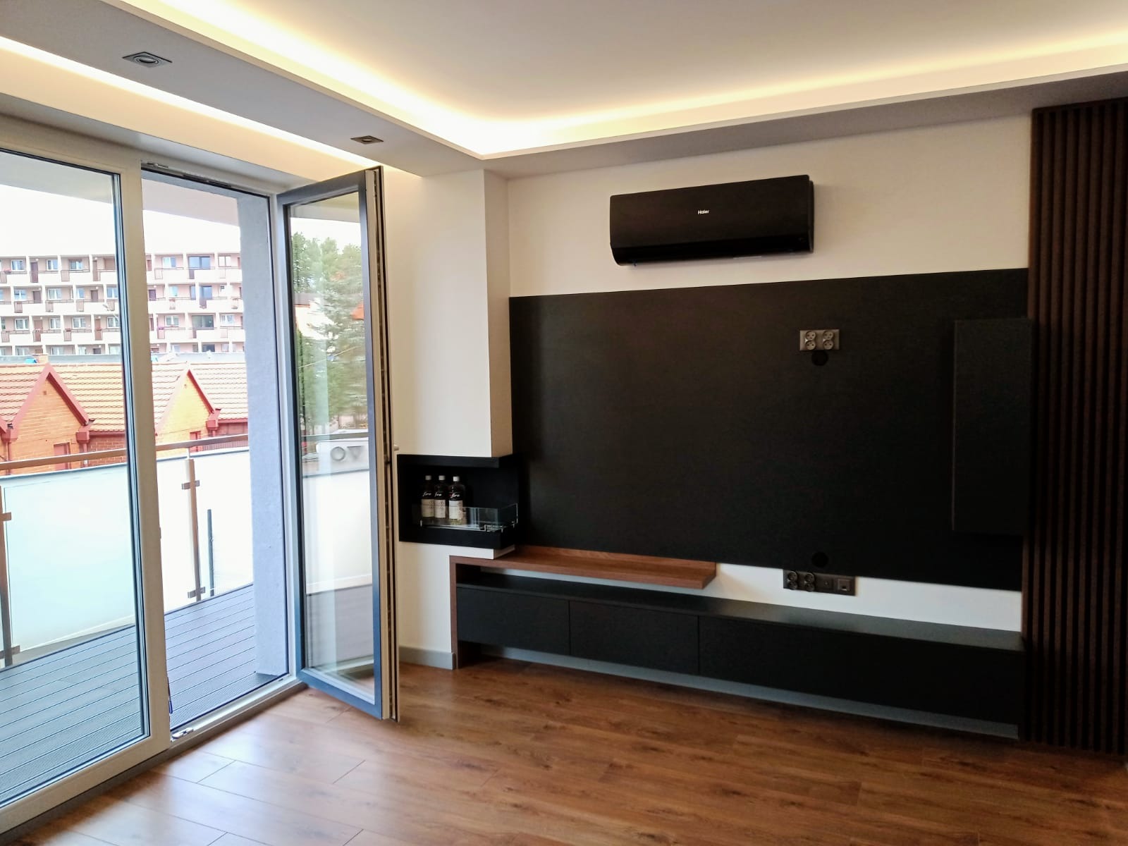 Nowoczesny salon z otwartym balkonem, drewnianą podłogą i czarną ścianą telewizyjną z minimalistyczną szafką RTV i półką na alkohol, oświetlony listwami LED na suficie.