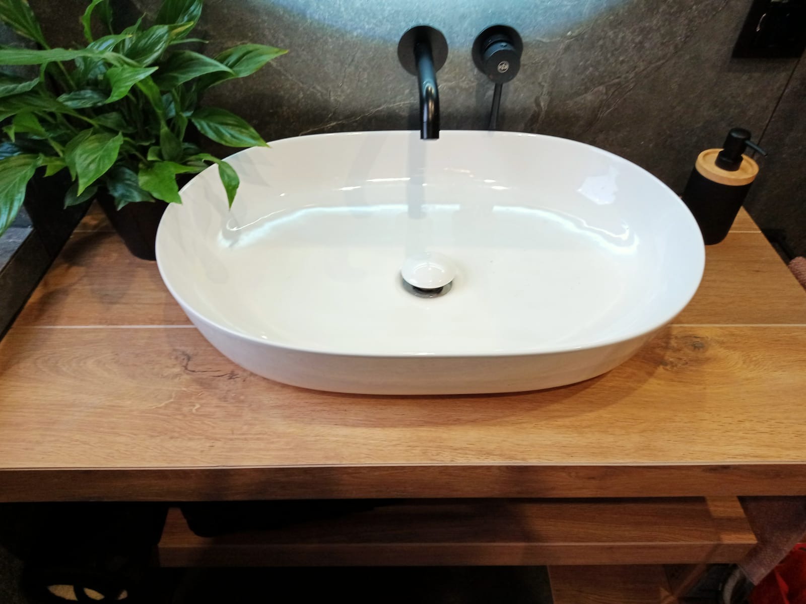 Biała, owalna umywalka nablatowa z czarną armaturą, drewnopodobny blat, szara ściana i zielona roślina doniczkowa w nowoczesnej łazience.