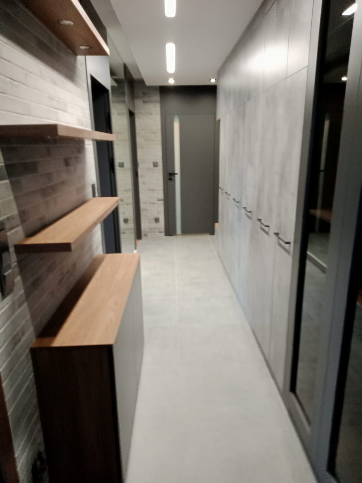 Wnętrze korytarza w Będzinie po remoncie, z zabudową szaf w szarym kolorze, drewnianymi półkami na ceglanej ścianie i szarą podłogą z płytek.