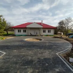 Szeroki podjazd z szarej kostki brukowej prowadzący do domu z czerwonym dachem, z okrągłym klombem pośrodku i białą obwódką, widok z perspektywy pieszego.