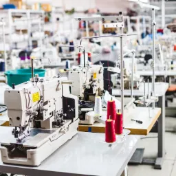 Rząd profesjonalnych maszyn do szycia w jasnej hali produkcyjnej, z widocznymi czerwonymi i białymi nićmi na szpulach, oraz półkami magazynowymi w tle.