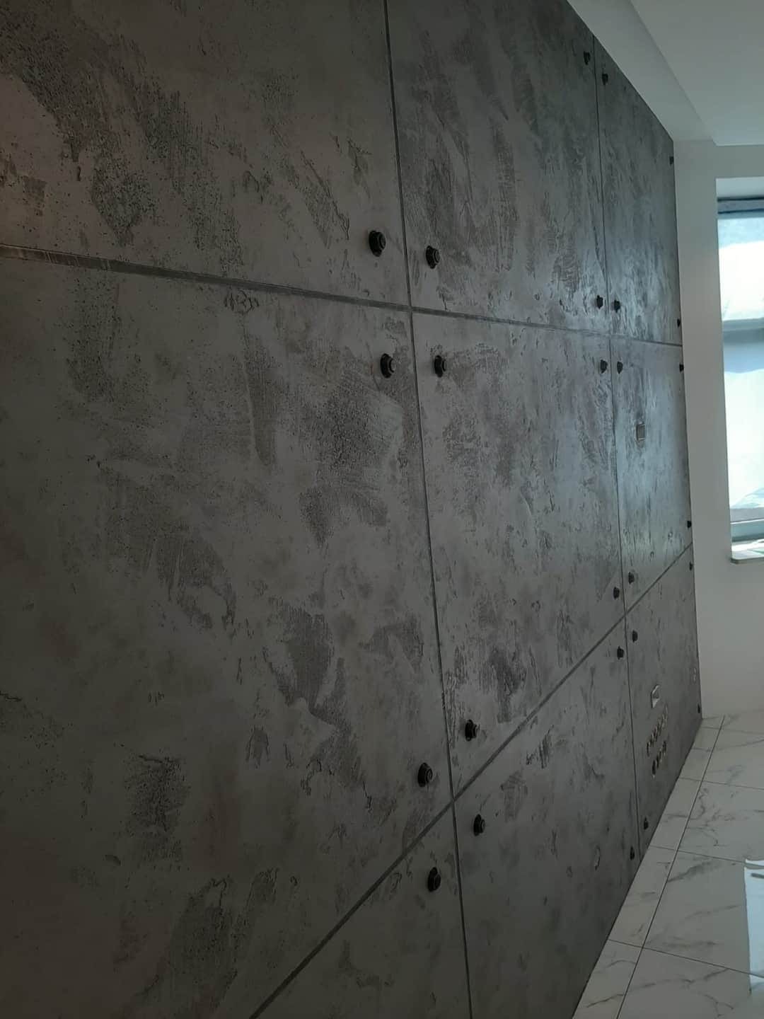 Ściana wykończona betonem architektonicznym w panelach, połączonych widocznymi śrubami, z marmurową podłogą i oknem w tle.