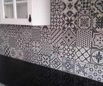 Ściana nad blatem kuchennym wyłożona różnorodnymi, wzorzystymi płytkami ceramicznymi w odcieniach szarości i czerni, z fragmentem białej szafki wiszącej z czarnym uchwytem.
