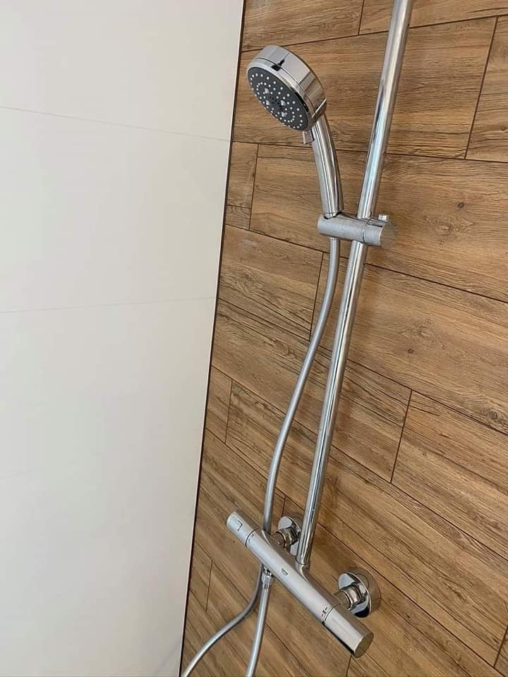 Bateria prysznicowa z chromowaną słuchawką na ścianie z paneli imitujących drewno, obok białej ściany.