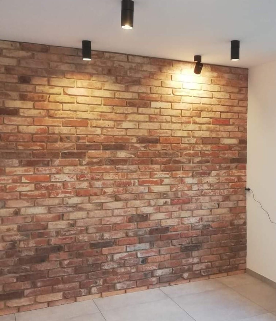 Ściana z dekoracyjnej cegły w odcieniach czerwieni i brązu, oświetlona punktowymi lampami sufitowymi w nowoczesnym wnętrzu z jasną podłogą.