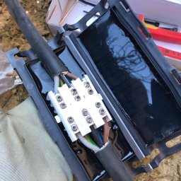 MMASS Michał Szot - Zalewanie żywicą złączki kablowej niskiego napięcia w czarnej obudowie z widocznymi przewodami i kostką przyłączeniową, otoczenie piaszczyste.