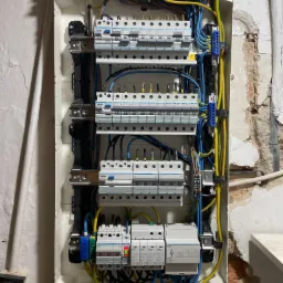 Otwarta skrzynka rozdzielcza z zamontowanymi bezpiecznikami i okablowaniem wewnątrz, widoczne przewody w kolorach niebieskim, żółtym i zielonym. Ściana w tle z widocznymi śladami po skuciu tynku.