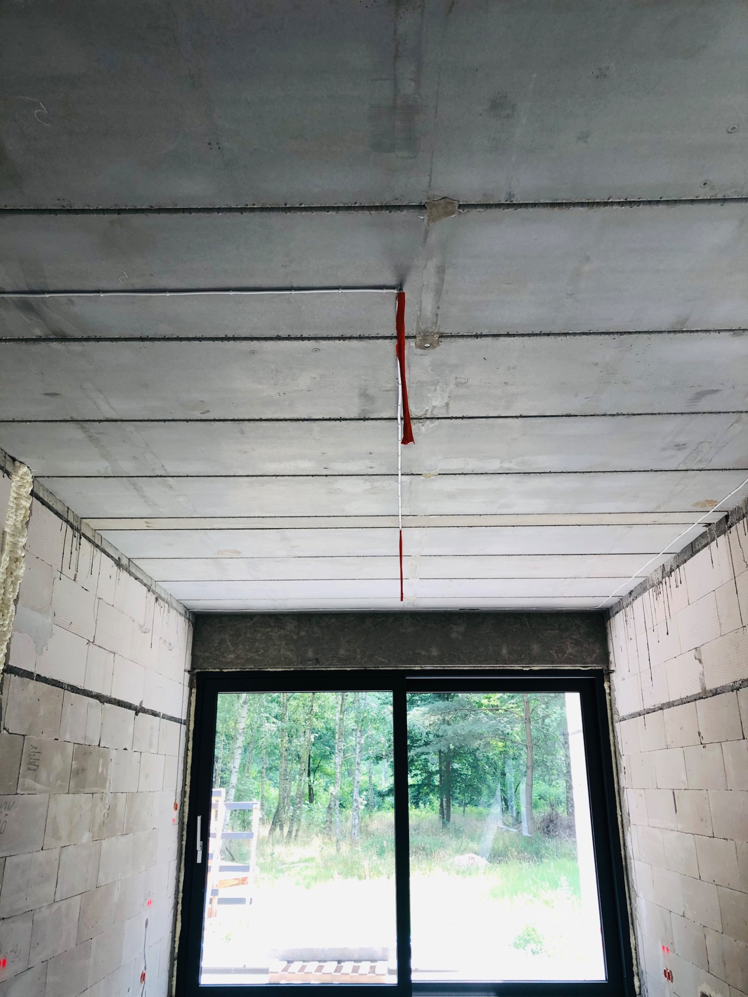 Surowy pokój w trakcie budowy z widocznymi przewodami elektrycznymi zwisającymi z betonowego stropu, ściany z białego betonu komórkowego, okno z widokiem na las.