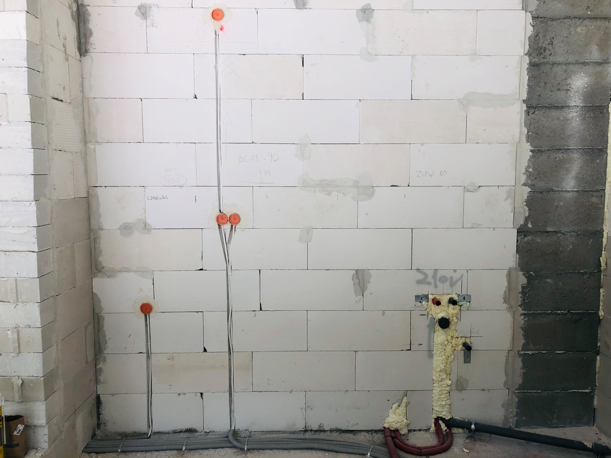 Wewnętrzna instalacja elektryczna w trakcie montażu w ścianie z białych bloczków, widoczne kable w peszlach i puszki instalacyjne, obok pion kanalizacyjny zabezpieczony pianką montażową.