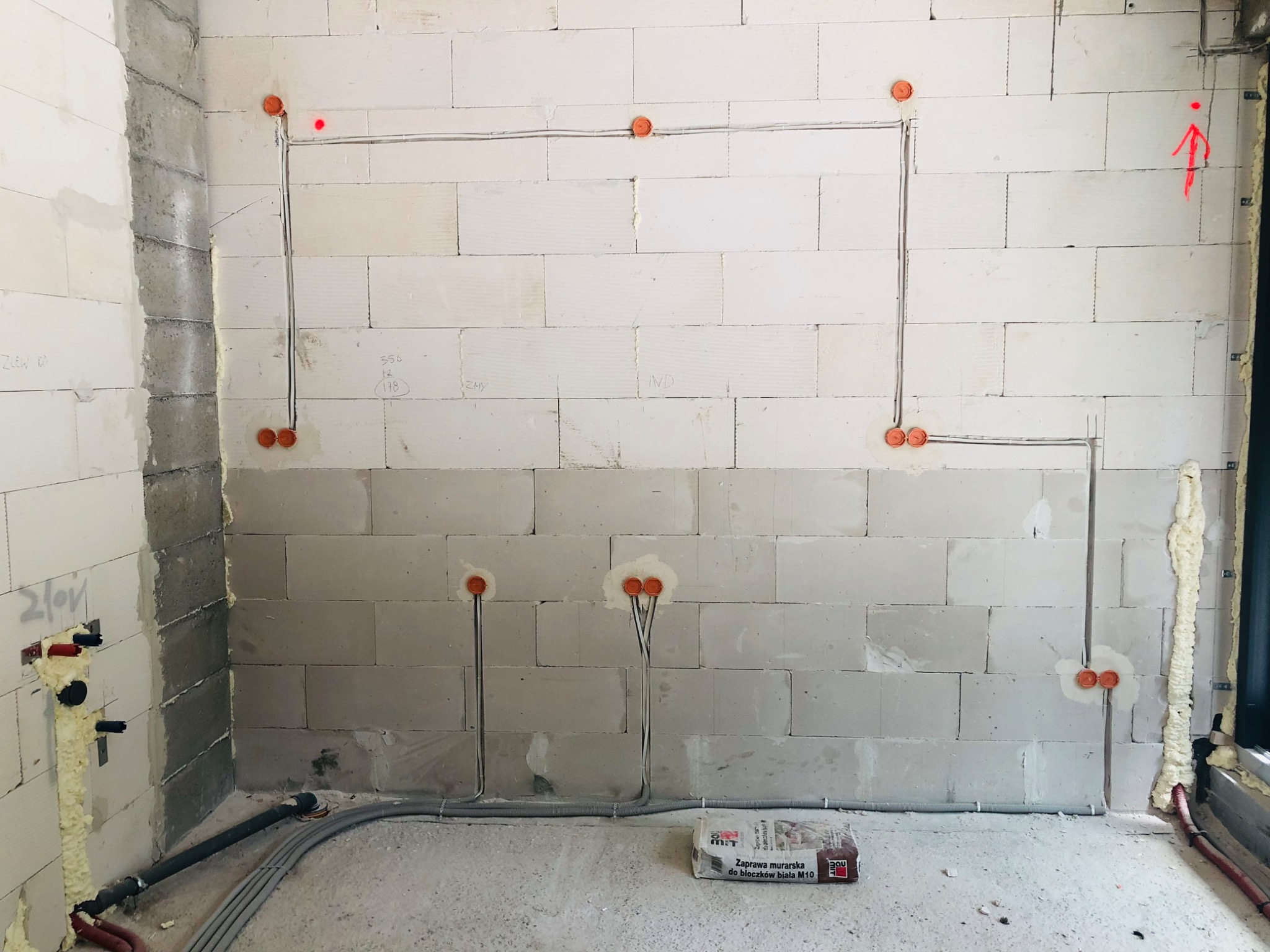 Surowy stan budowy: instalacja elektryczna w ścianach z betonu komórkowego, widoczne kable w peszlach i puszki instalacyjne, zaprawa murarska na podłodze.