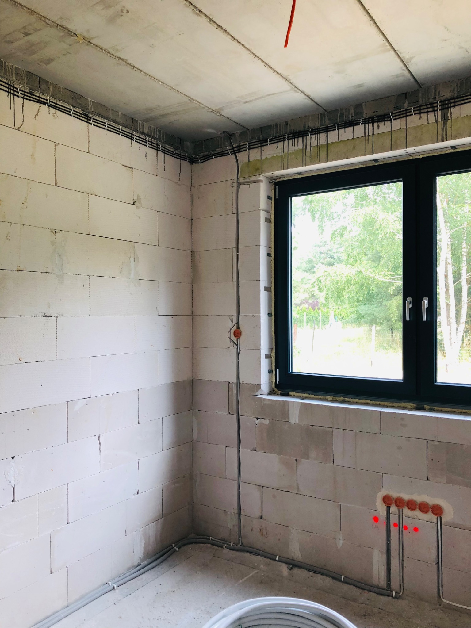 Surowy pokój w trakcie budowy z widocznymi przewodami elektrycznymi w ścianach z betonu komórkowego i pod sufitem, oraz oknem z widokiem na drzewa.