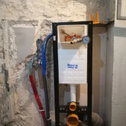 Stelaż podtynkowy WC Roca zamontowany na tle surowej, częściowo otynkowanej ściany z widocznymi rurami doprowadzającymi wodę.