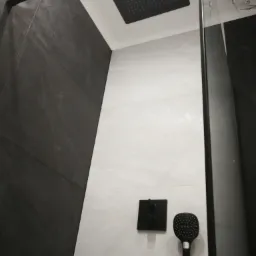 Nowoczesna kabina prysznicowa z czarną armaturą: deszczownica sufitowa i słuchawka prysznicowa na tle szarych płytek.