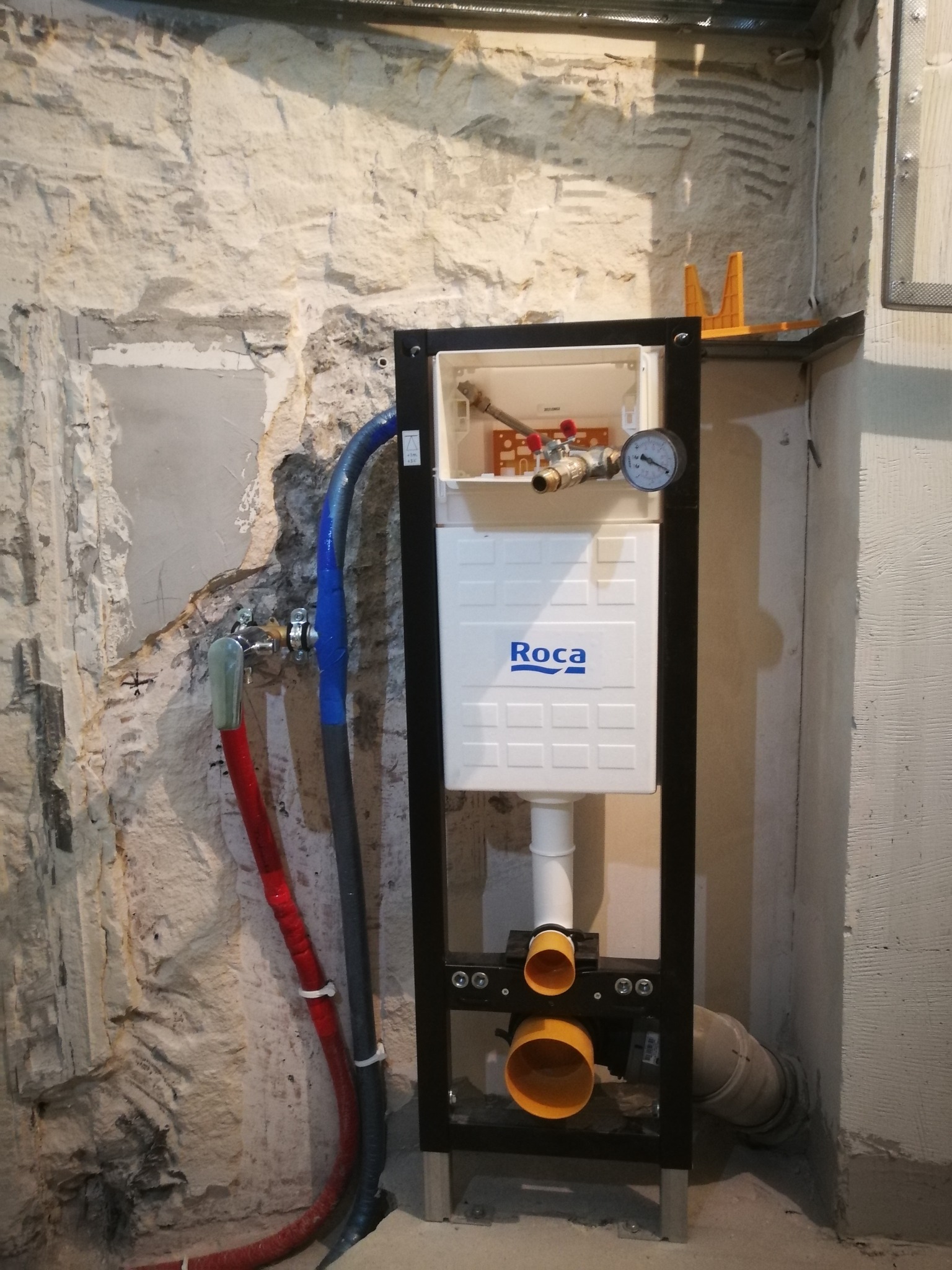 Stelaż podtynkowy WC Roca zamontowany na tle surowej, częściowo otynkowanej ściany z widocznymi rurami doprowadzającymi wodę.