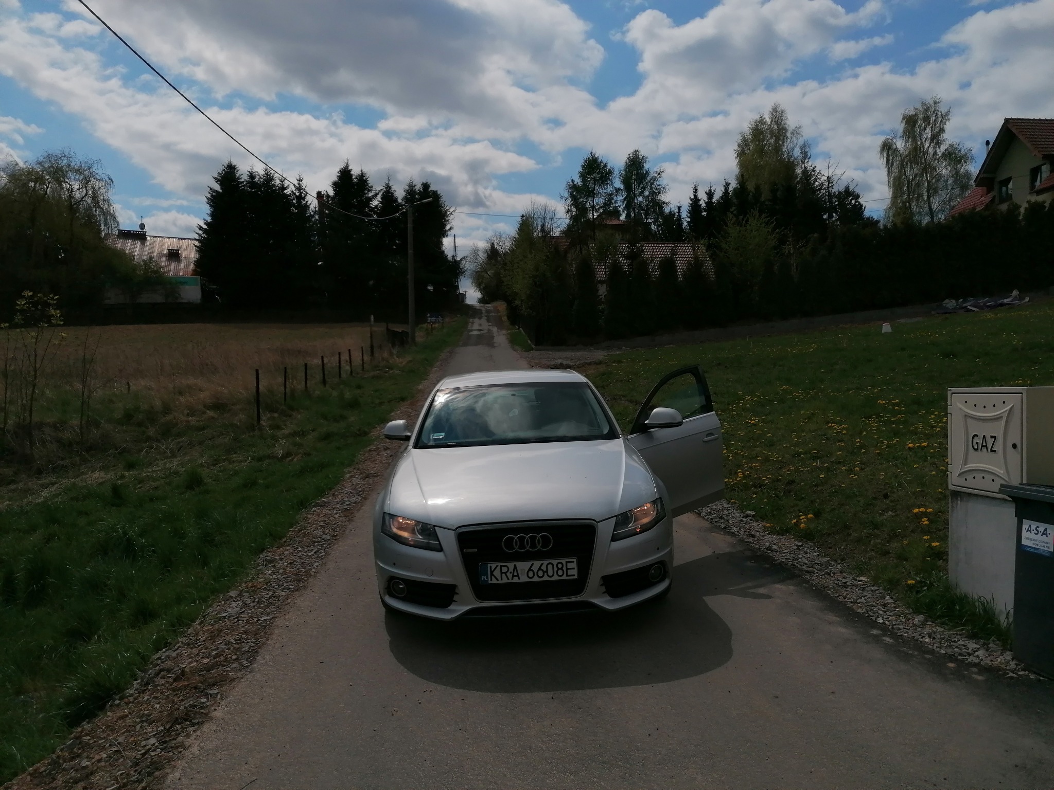 Srebrny samochód osobowy marki Audi z otwartymi drzwiami kierowcy, zaparkowany na asfaltowej drodze w wiejskiej scenerii z polami i drzewami w tle, widok z przodu.