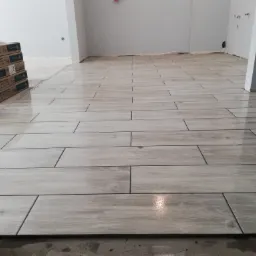 Ułożona podłoga z podłużnych, jasnych płytek imitujących drewno, widoczne paczki płytek i narzędzia glazurnicze w tle, surowy stan wykończenia ścian.