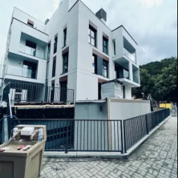 Nowoczesny budynek apartamentowy z białymi elewacjami i ciemnymi oknami, otoczony metalowym ogrodzeniem, widoczne prace wykończeniowe na zewnątrz.