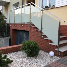 Zewnętrzne schody z balustradą ze stali nierdzewnej i mlecznego szkła, prowadzące do drzwi wejściowych budynku z numerem 10, otoczone dekoracyjnym żwirem i niską roślinnością.