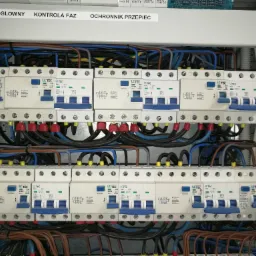 Otwarta skrzynka elektryczna z widocznymi bezpiecznikami, przewodami i oznaczeniami 'Wył. Główny', 'Kontrola Faz' oraz 'Ochronnik Przepięć'.