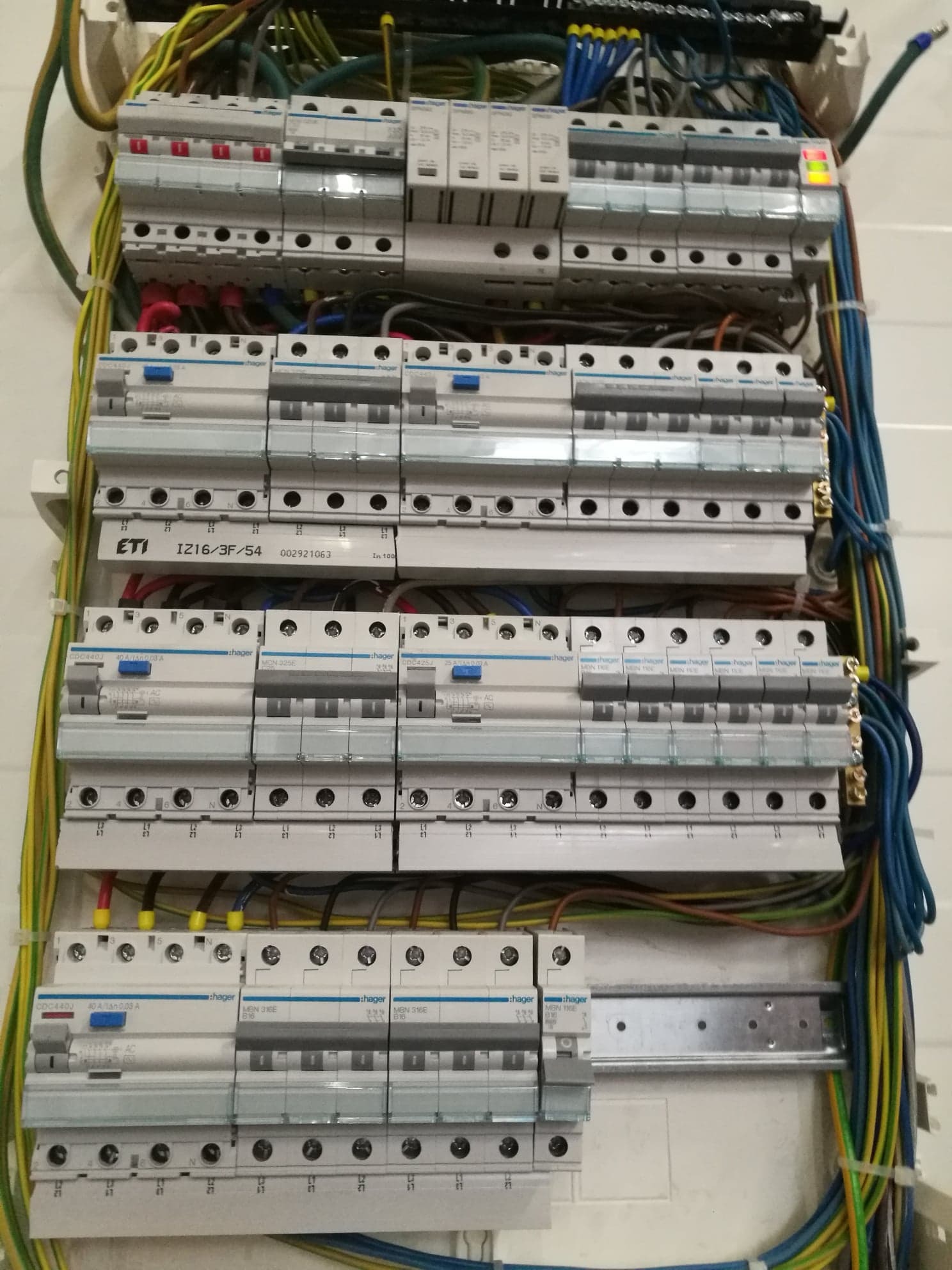 Rozdzielnica elektryczna z zainstalowanymi bezpiecznikami ETI i Hager, widoczne przewody w różnych kolorach, niektóre z oznaczeniami, zamontowana na białej ścianie.