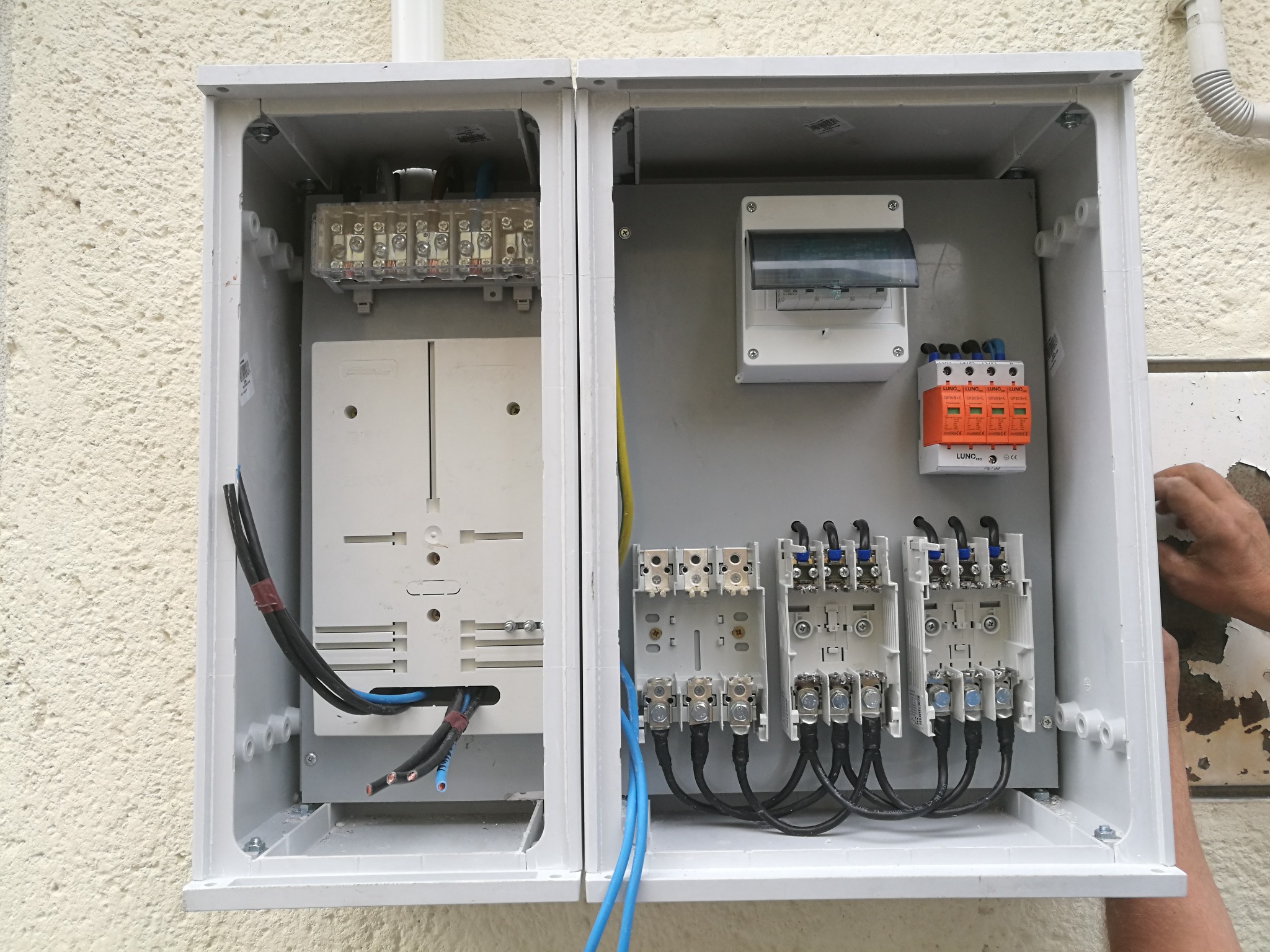 Otwarta, dwuczęściowa skrzynka elektryczna z widocznymi bezpiecznikami, listwami zaciskowymi, przewodami i ogranicznikiem przepięć, zamontowana na elewacji budynku. Widoczne ręce podczas pracy.