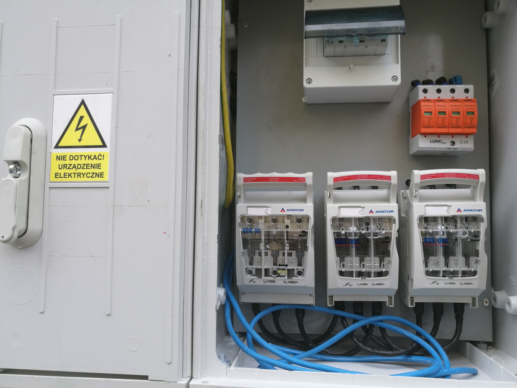 Otwarta skrzynka elektryczna z bezpiecznikami Apator RBK 00, zabezpieczeniem przeciwprzepięciowym LUNG PRO, niebieskimi przewodami i żółtą naklejką ostrzegawczą 'Nie dotykać! Urządzenie elektryczne'.