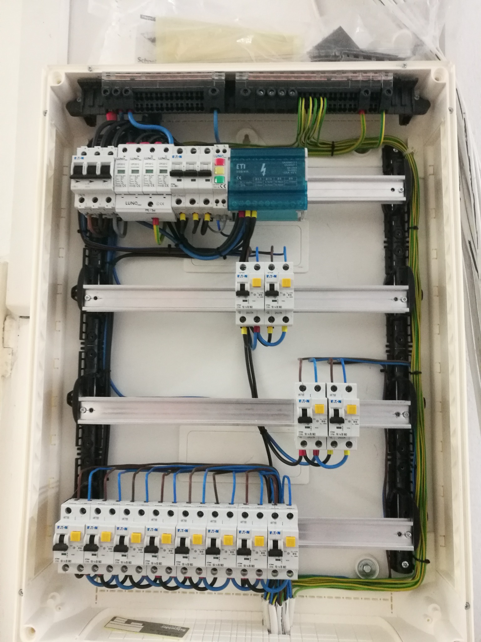 Otwarta skrzynka elektryczna z zainstalowanymi bezpiecznikami, modułem ochronnym i okablowaniem, widoczne elementy marek LUNO i ETI, częściowo zakryta folią ochronną w górnym rogu.