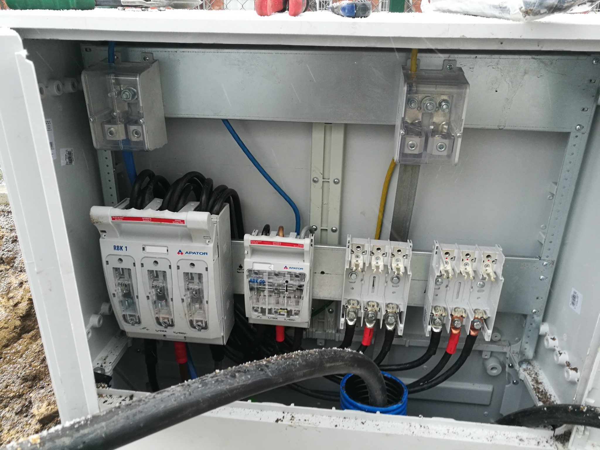 Rozdzielnia elektryczna z bezpiecznikami i przewodami w obudowie, widoczne elementy instalacji, w tym zabezpieczenia i złącza.