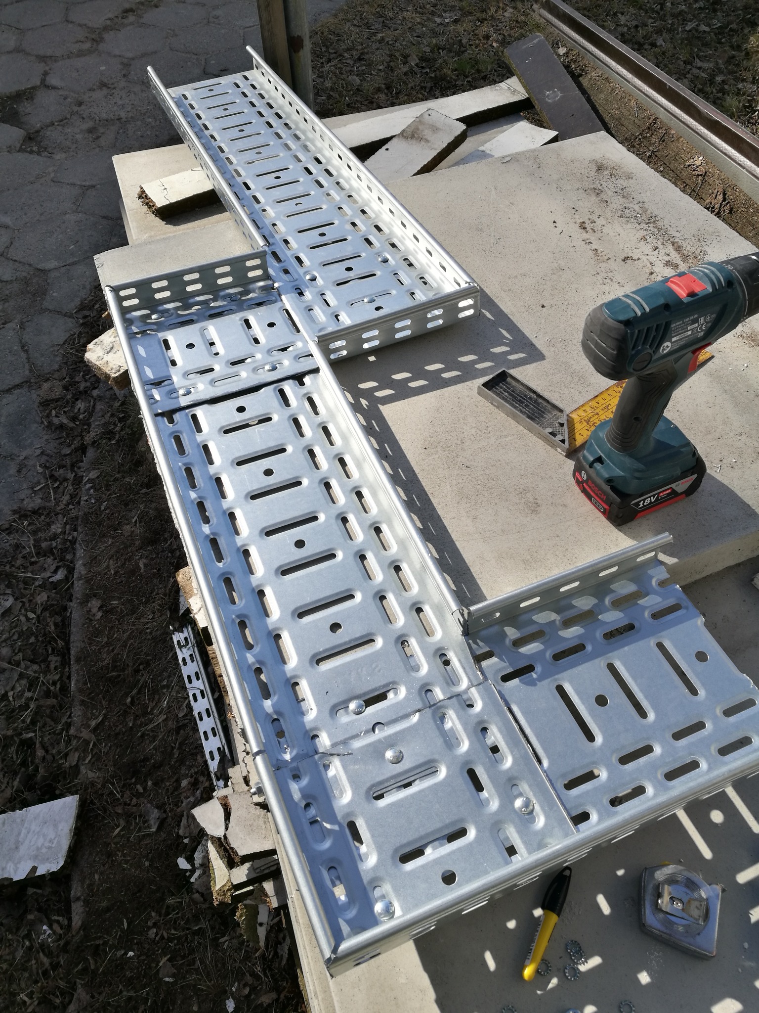 Dwie metalowe korytka kablowe z perforacją, ułożone na betonowych płytach, obok wiertarka akumulatorowa, miarka zwijana i marker.