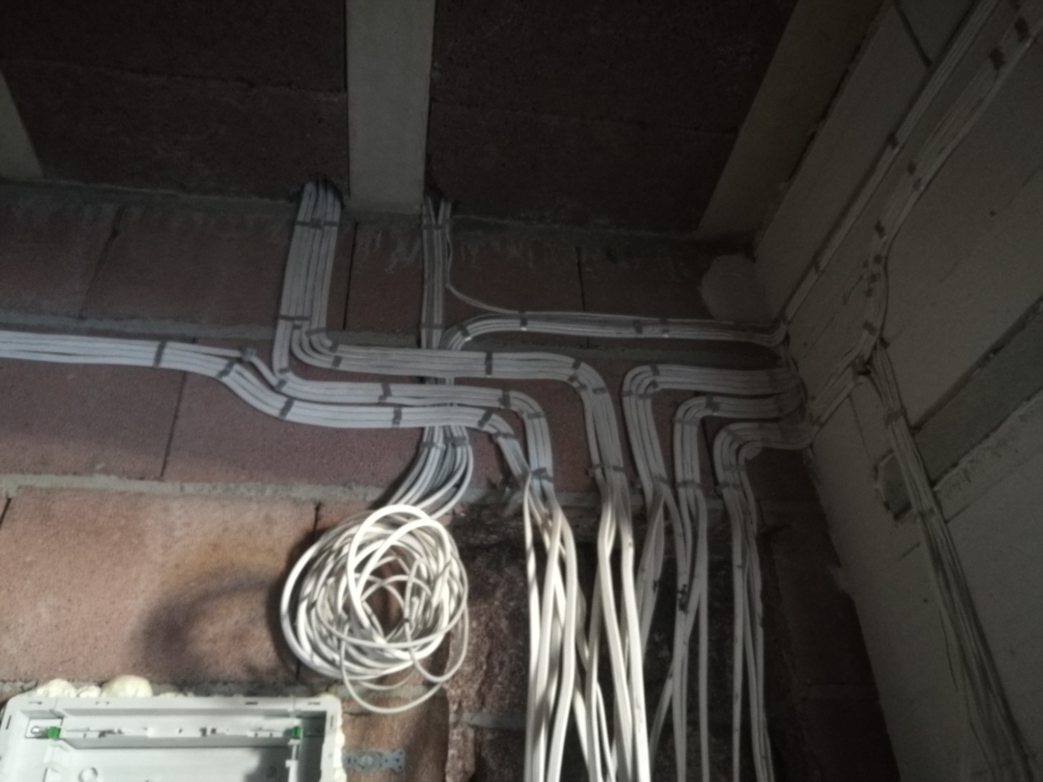 Ściana z cegły z poprowadzonymi kablami elektrycznymi w peszlach, część zwinięta w pętlę, widoczna puszka instalacyjna z pianką montażową.