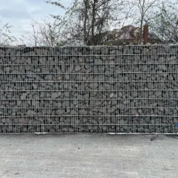 Gabion z szarego kamienia w metalowej siatce tworzy ogrodzenie. Widoczne drzewa i dach domu w tle. Na dole kostka brukowa i zwój niebieskiego kabla.