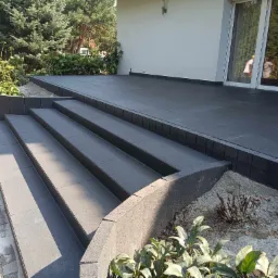 Nowoczesny taras z ciemnoszarej kostki, schody z tego samego materiału. Widoczne detale wykończenia i zieleń w tle. Ujęcie z niskiej perspektywy.