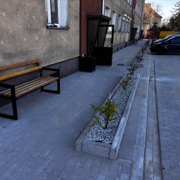 Brukarstwo Krzysztof Ciesielski - Szara kostka brukowa przed budynkiem z ławką i donicami z roślinami. W tle zaparkowane samochody i drzewa.