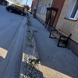 Brukarstwo Krzysztof Ciesielski - Ułożona szara kostka brukowa tworzy chodnik wzdłuż budynku z ławkami i rabatą z białym żwirem i roślinami. W tle zaparkowane samochody i błękitne niebo.
