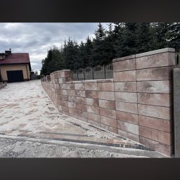 Brukarstwo Krzysztof Ciesielski - Mur z kostki brukowej w odcieniach brązu, oddzielający podjazd od trawnika, z widocznym domem i drzewami w tle. Ujęcie z niskiej perspektywy podkreśla strukturę muru.