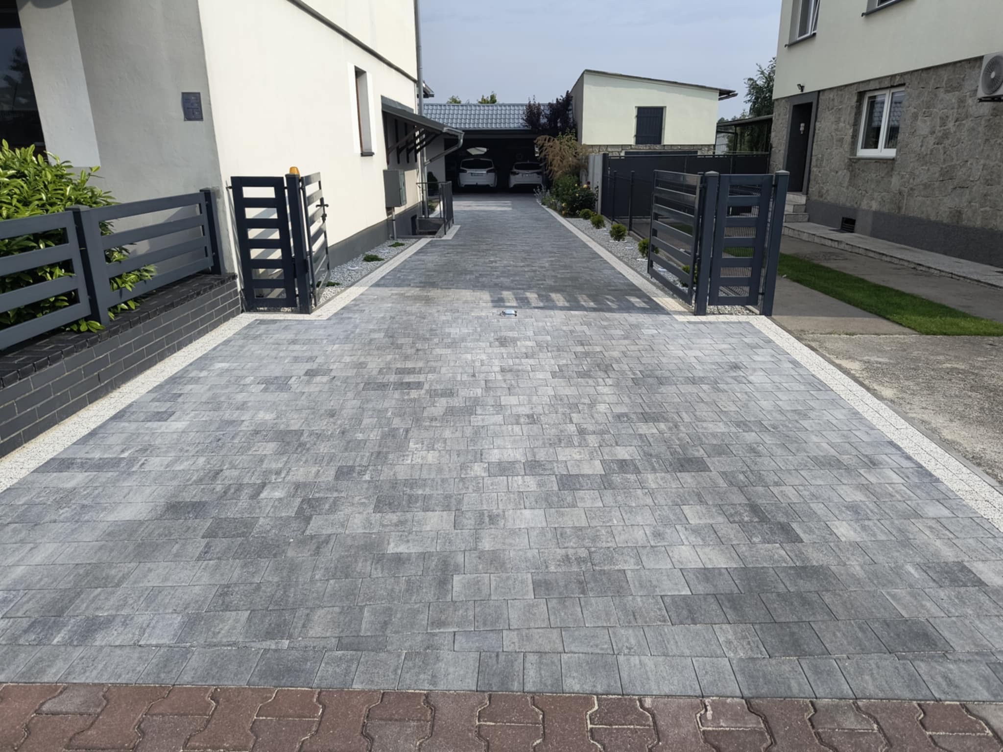 Szara kostka brukowa na podjeździe, obrzeża z drobnych kamyczków, nowoczesne ogrodzenie, dom z białymi ścianami w tle, widok na garaż.