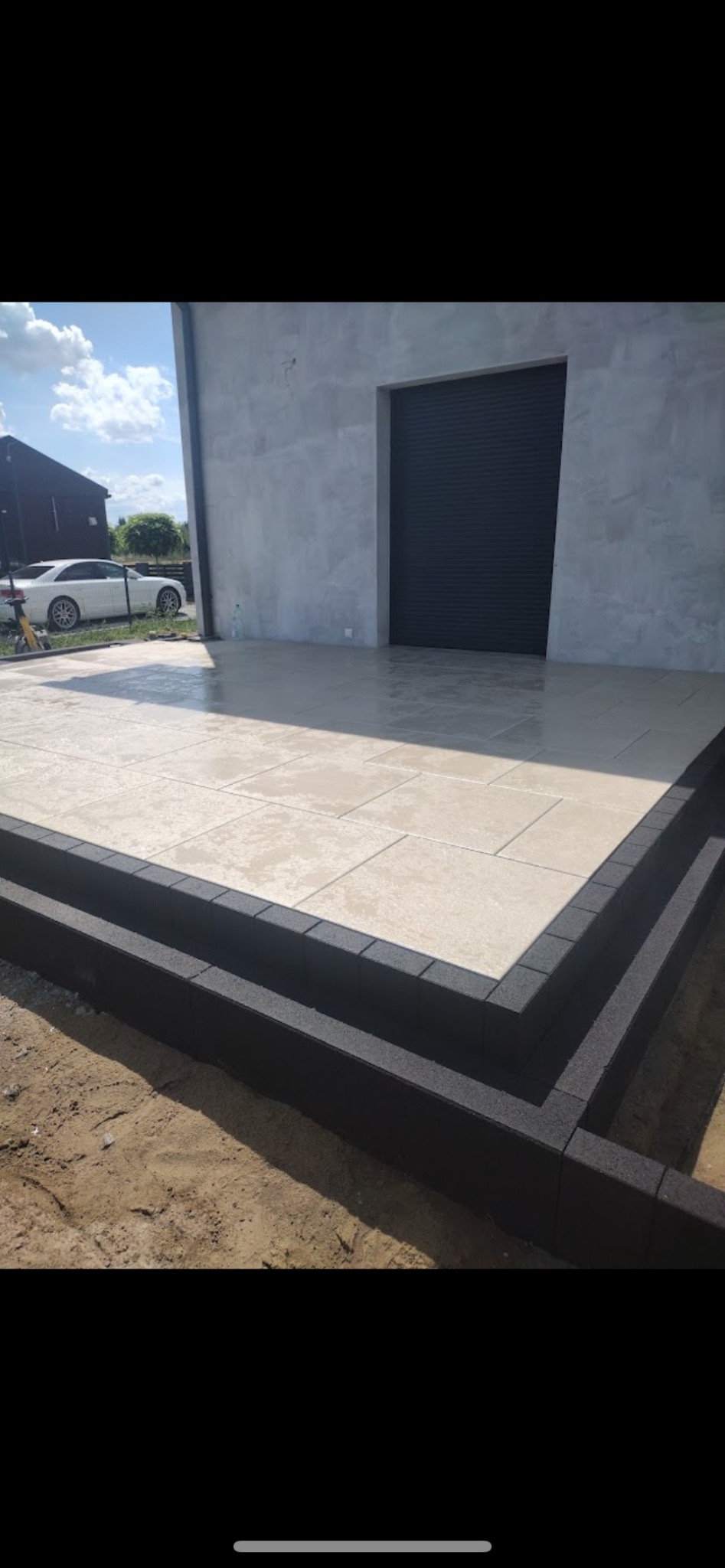 Jasne płytki tarasowe z ciemnymi obrzeżami z kostki, przylegające do budynku z szarą bramą garażową. W tle biały samochód i budynek w budowie.